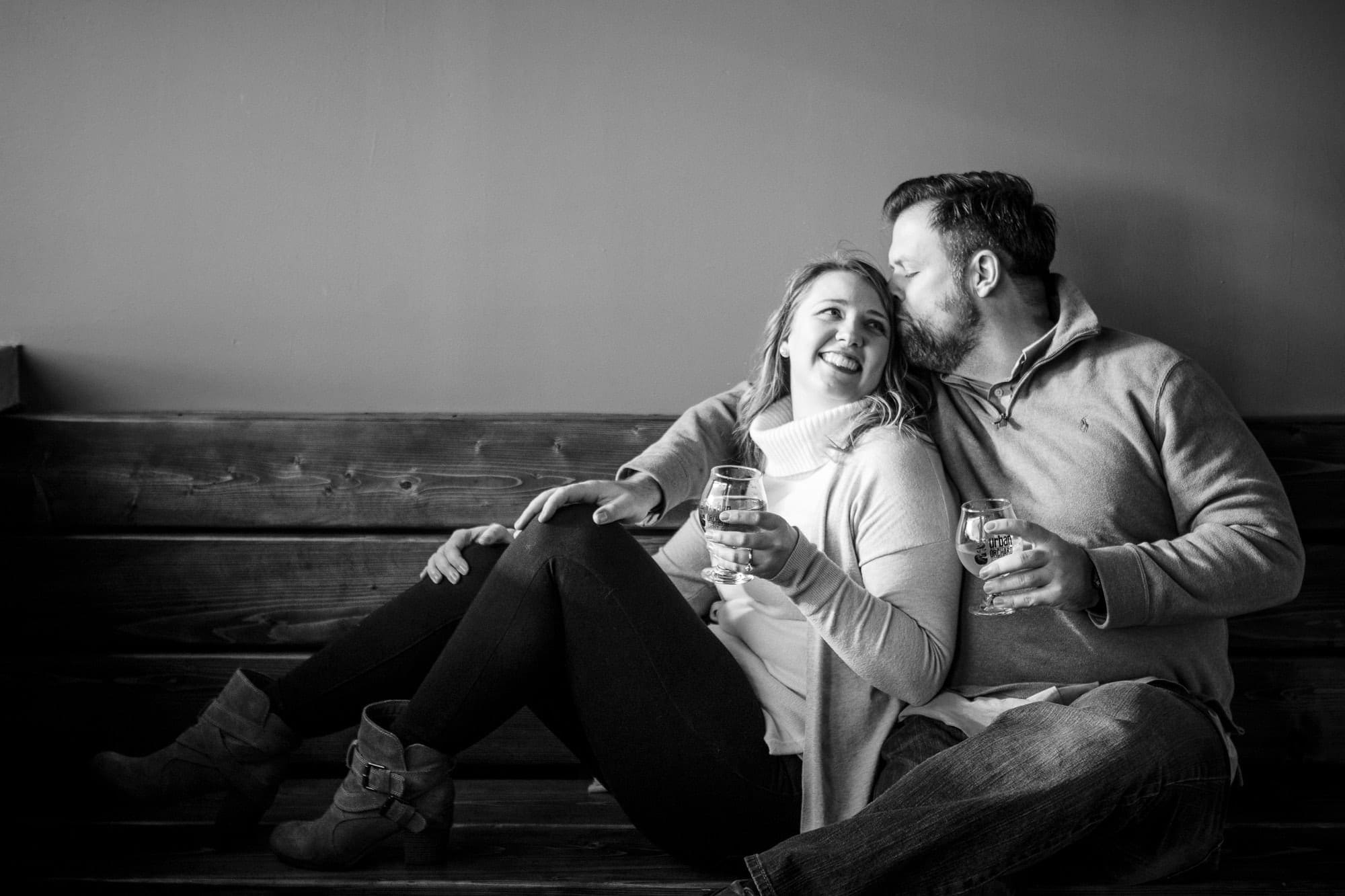 Black and white photo of man kissing fiance on the forehead while sitting on bench in restaurant drinking wine in Downtown Asheville North Carolina photography done by Kathy Beaver.
