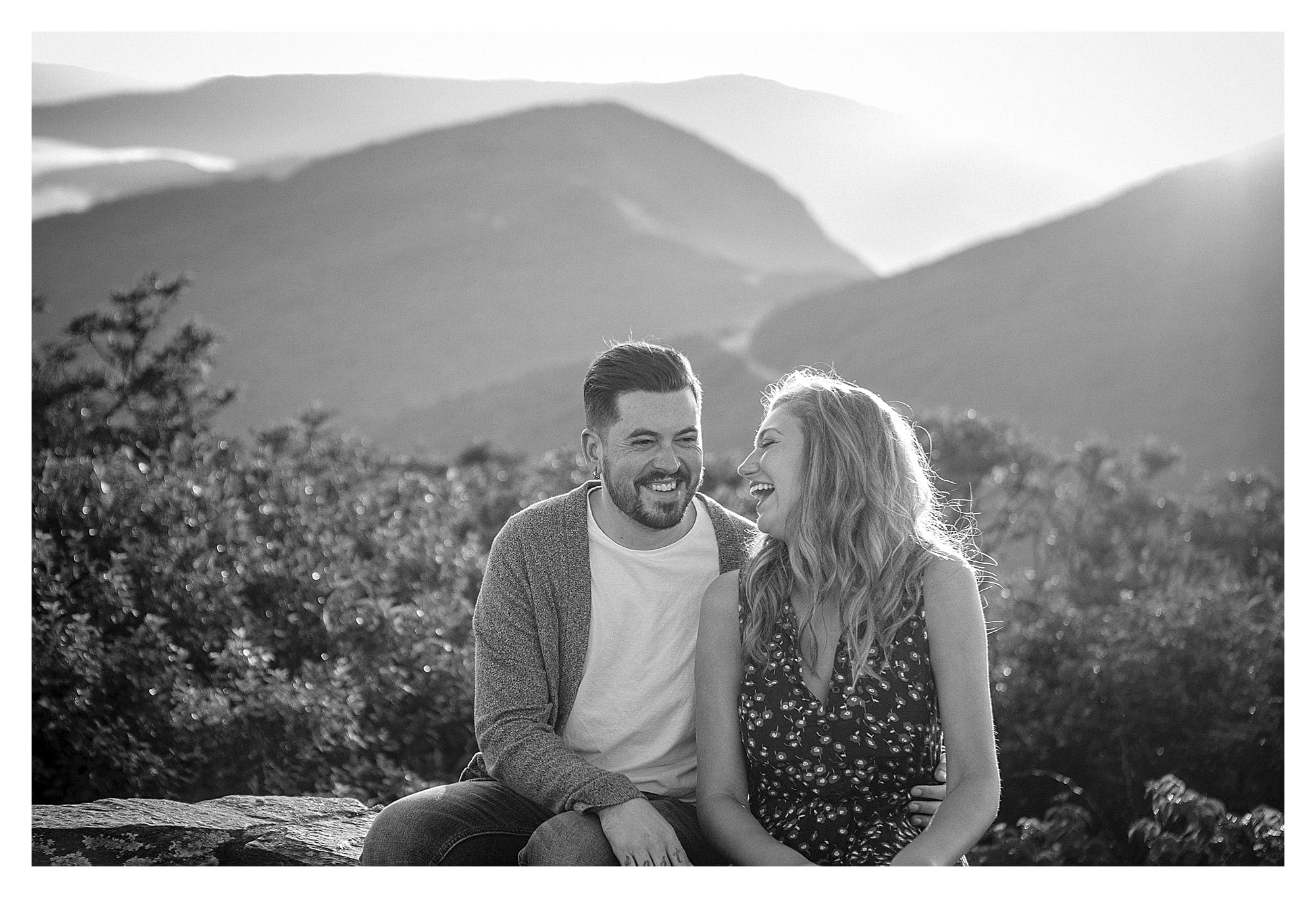 Black and white photo of engaged couple sitting and laughing with mountains behing them