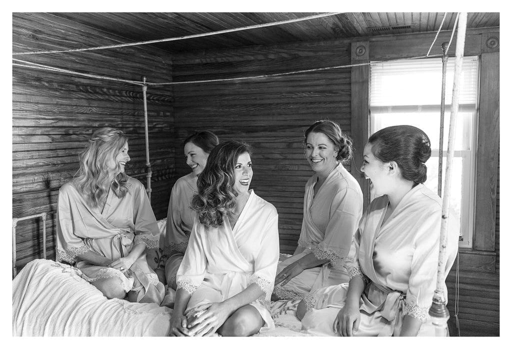 black and white image of bride and bridesmaids sitting on bed laughing while getting ready for wedding - kathy beaver photography