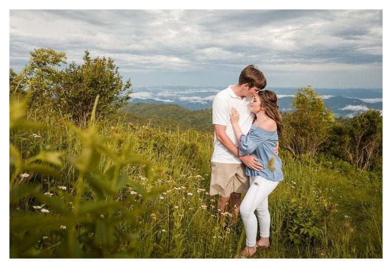 Young man kissing fiances forehead while standing in field with mountains in background