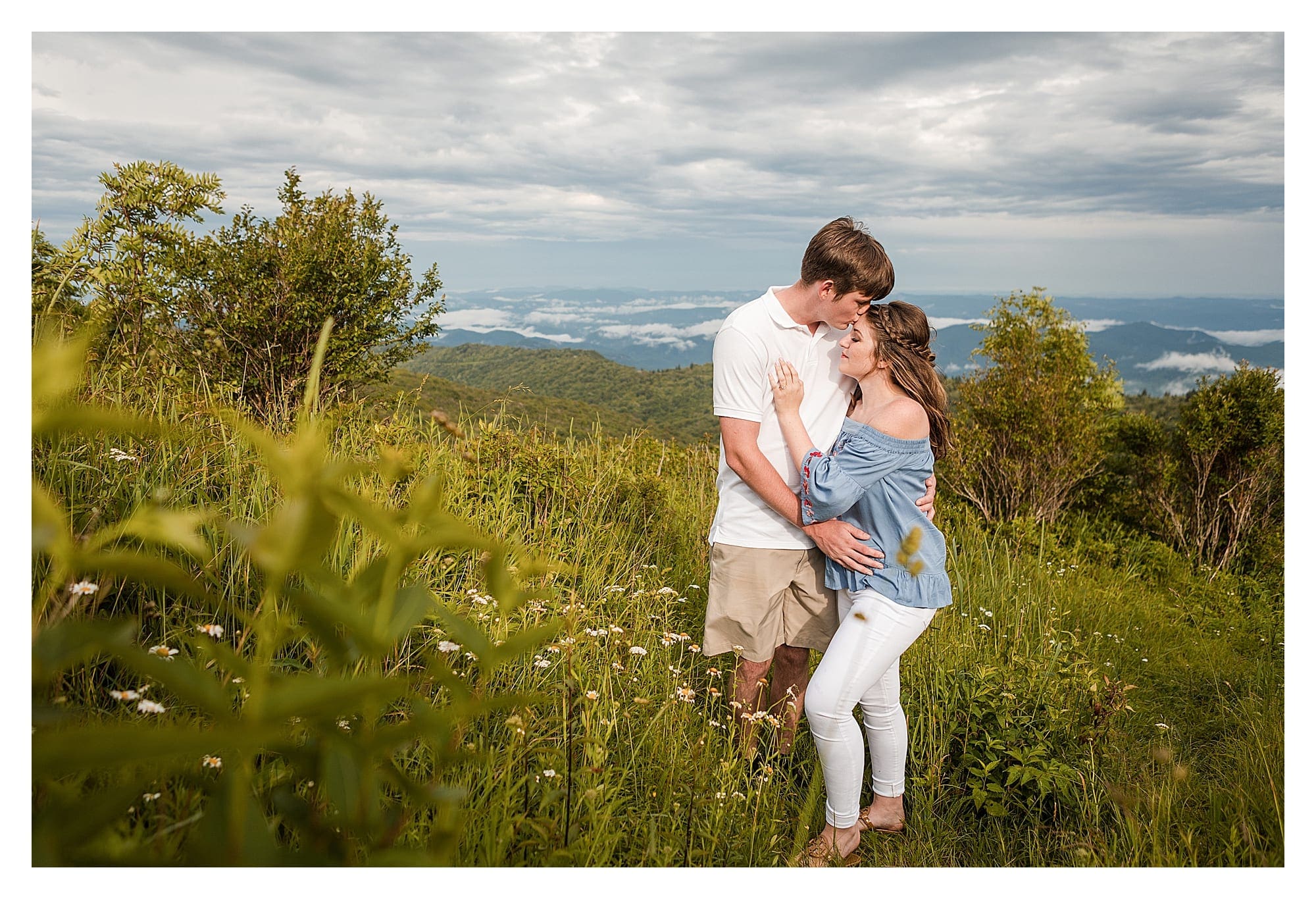 Young man kissing fiances forehead while standing in field with mountains in background