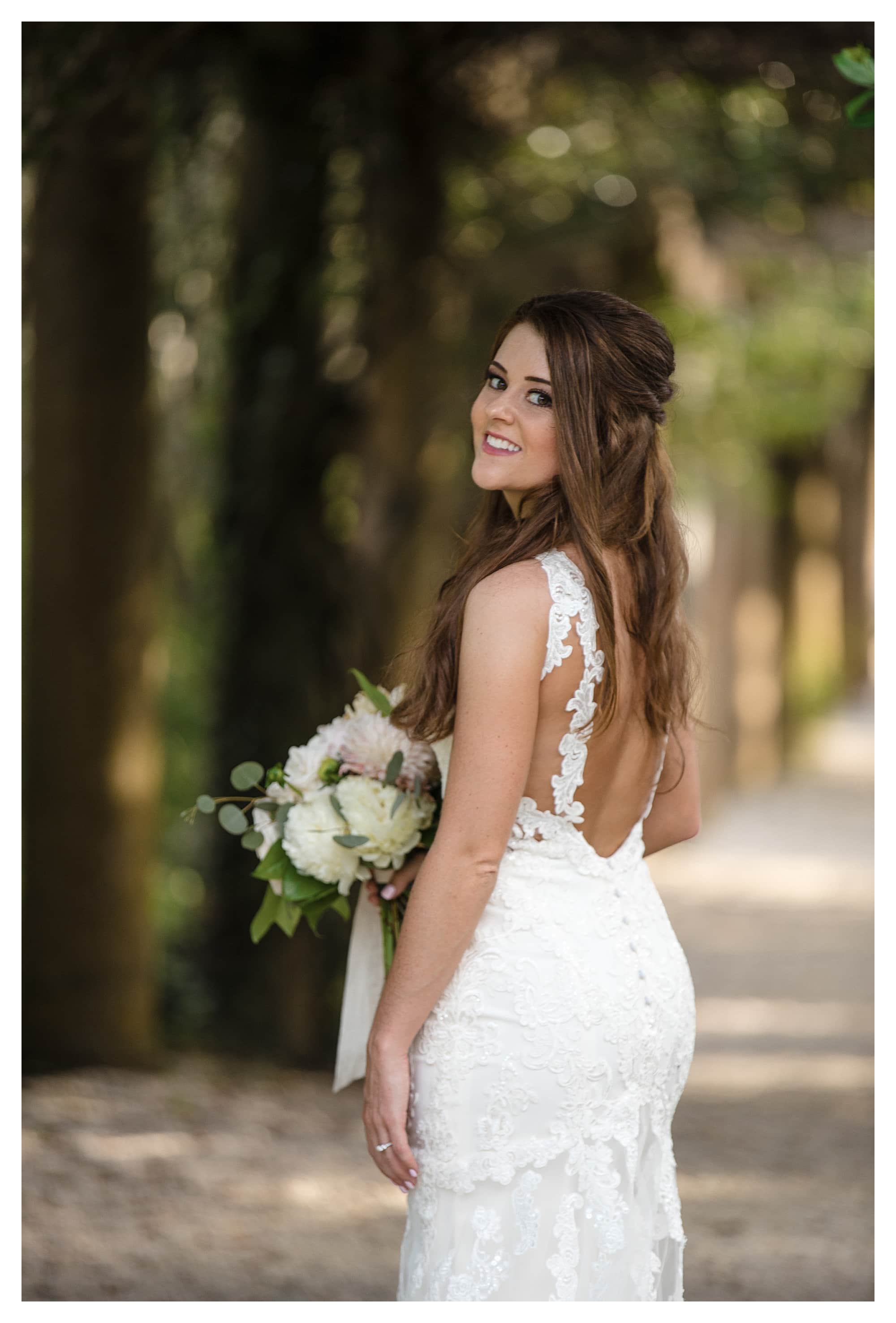 Close up of young bride smiling standing on treed pathway holding cream floral bouquet