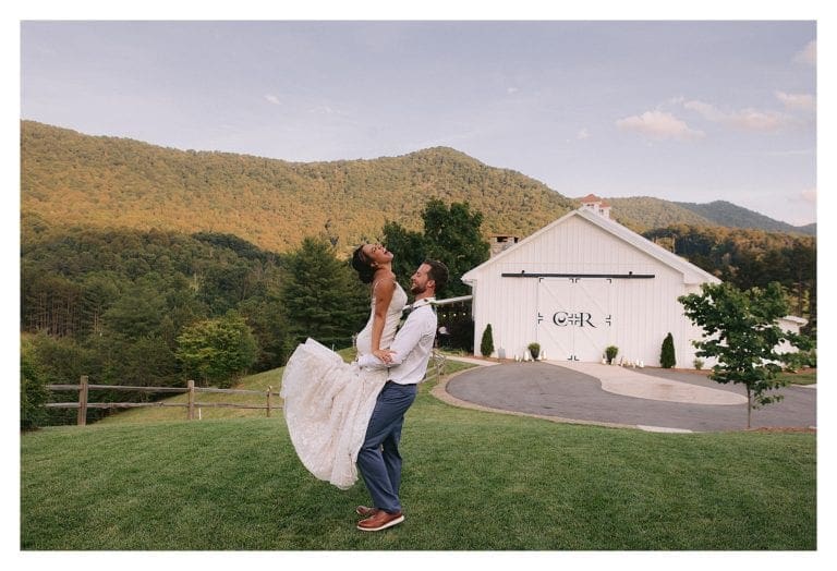 groom hugging bride and lifting her up in grassy field with mountains in background