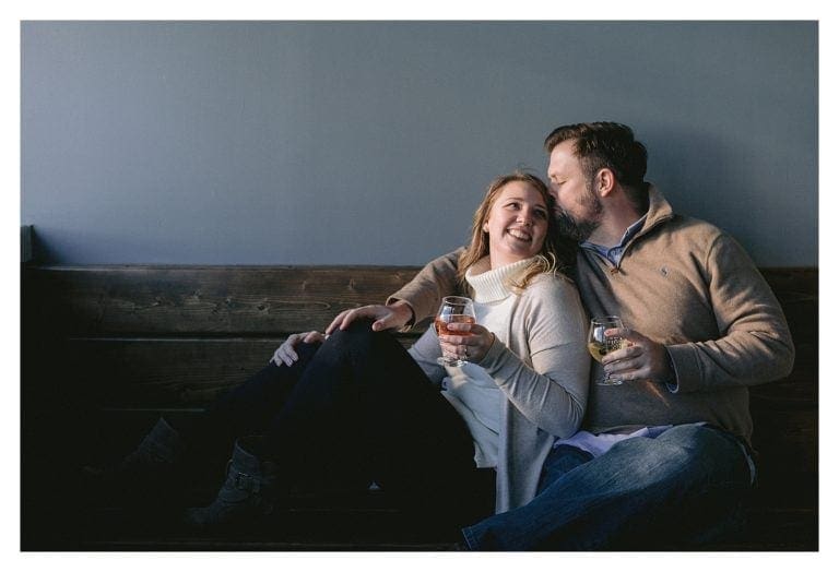 Young engaged couple sitting on wooden bench beside one another at urban orchard cidery in downtown Asheville, NC