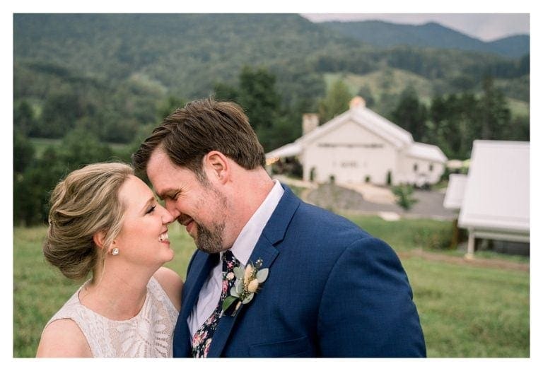 Bride and groom standing in field overlooking blue ridge mountains hugging and smiling at one another about to kiss