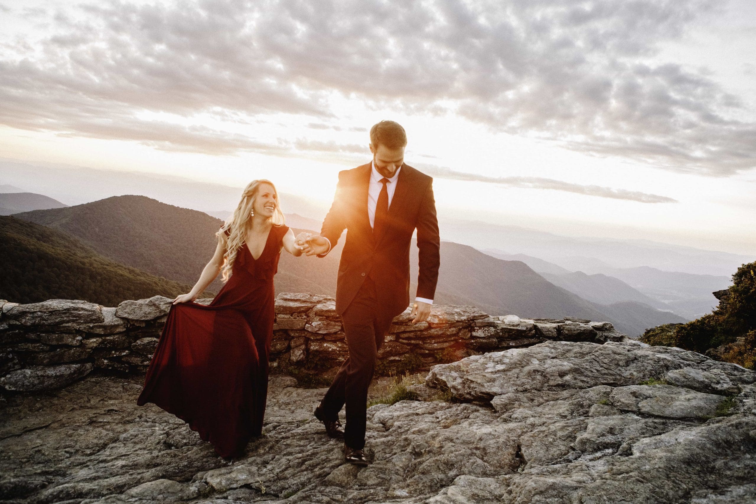 Man in suit holding fiances hand walking along rocky mountain side with mountain range in background at sunset