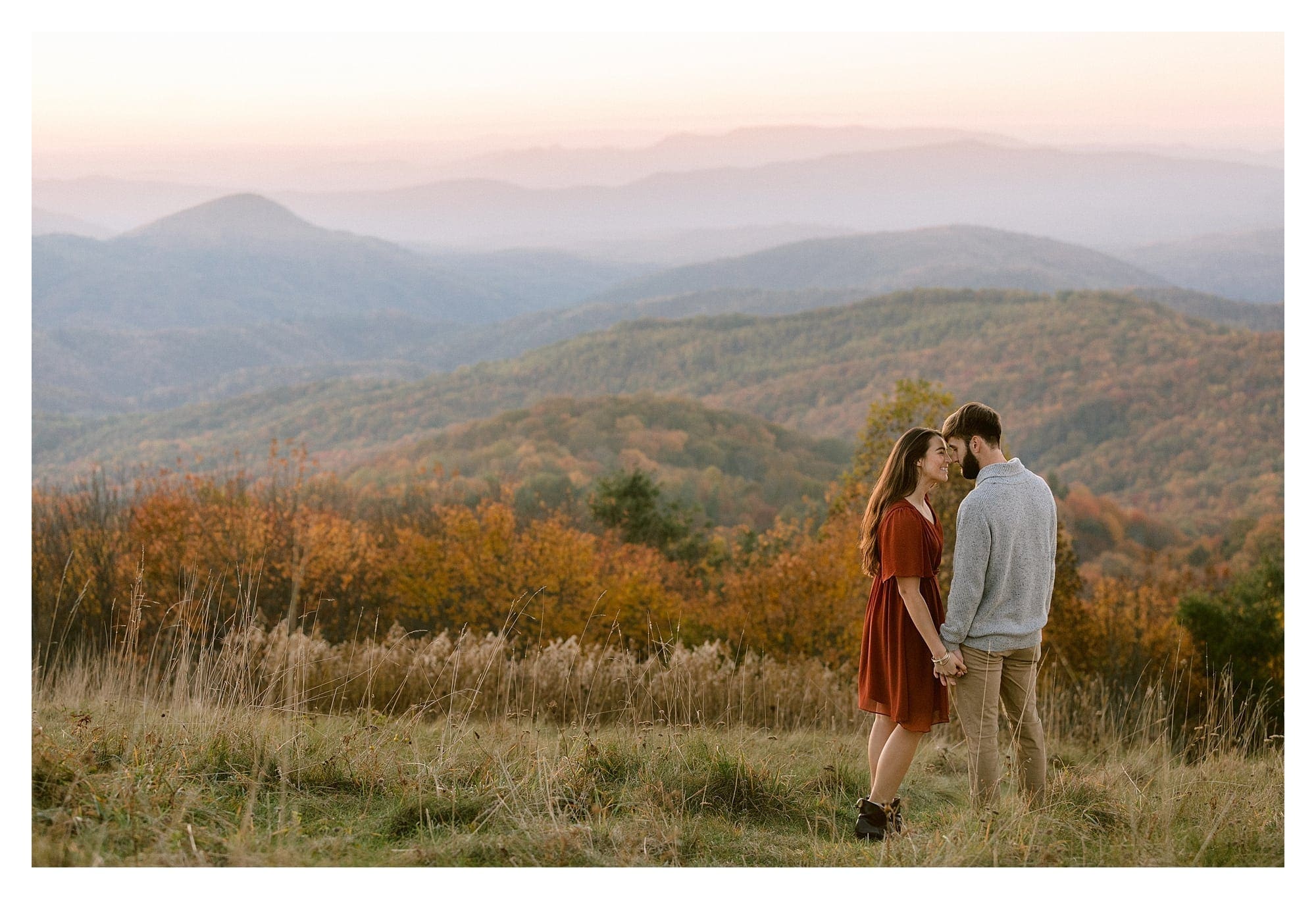 Young engaged couple kissing in golden grassy autumn field at sunset over looking mountain range