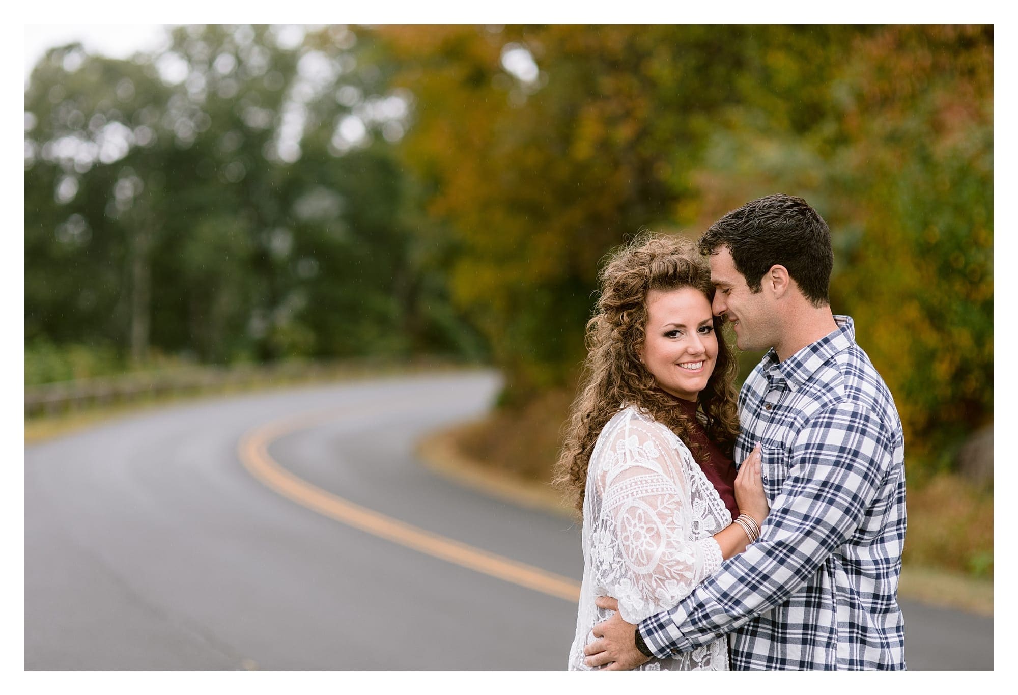 Engaged couple standing on road hugging one another and smiling with autumn trees behind them