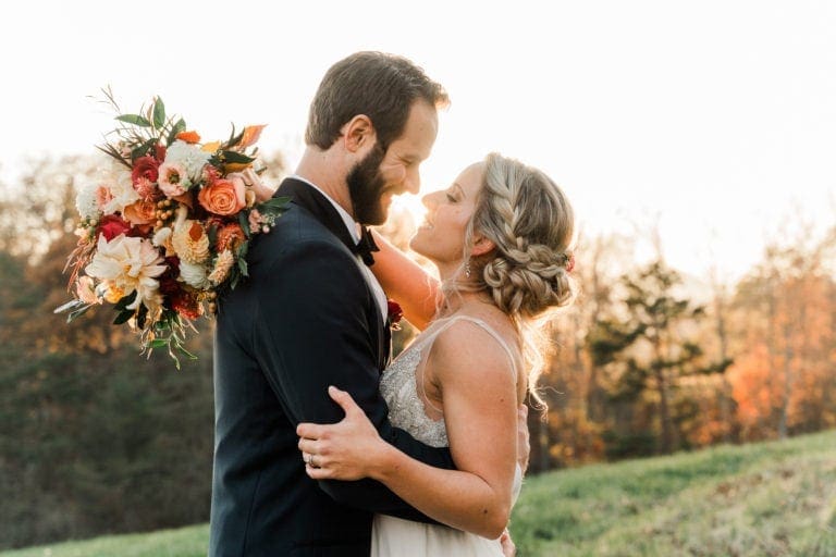 bride and groom standing on grassy field with sun setting behind them in the trees, arms around one another smiling about to kiss