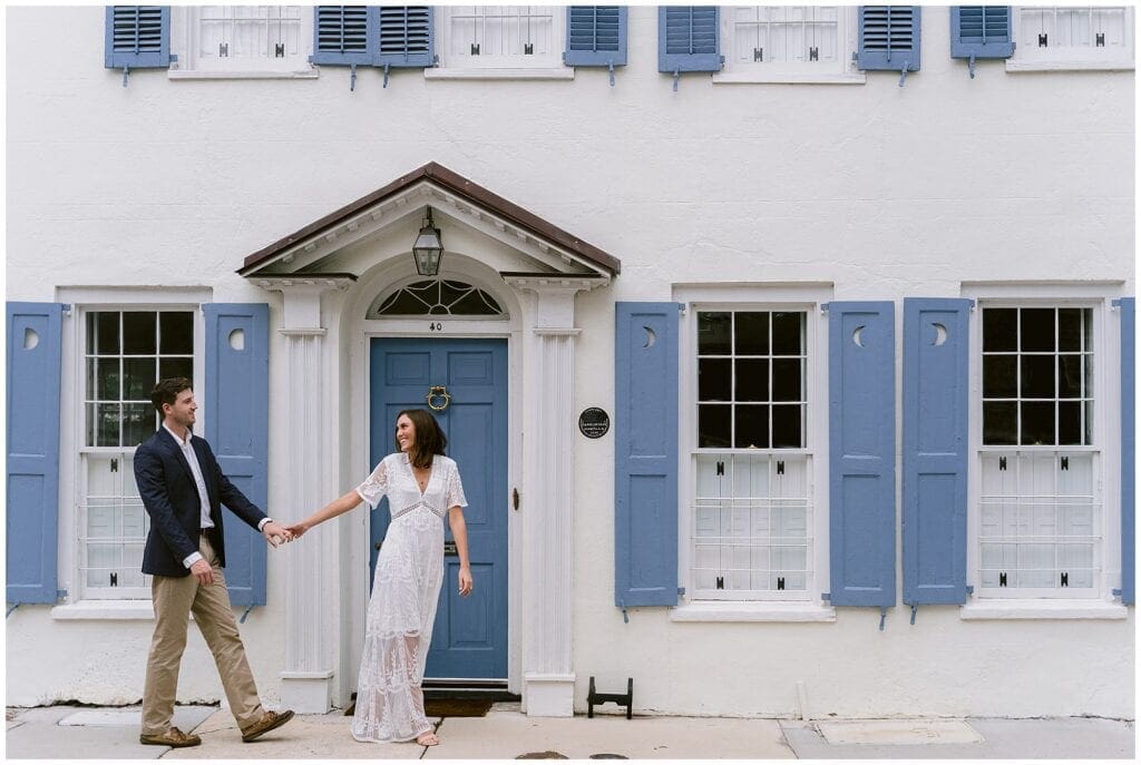 young engaged couple walking holding hands infront of white home with blue shutters in downtown charleston