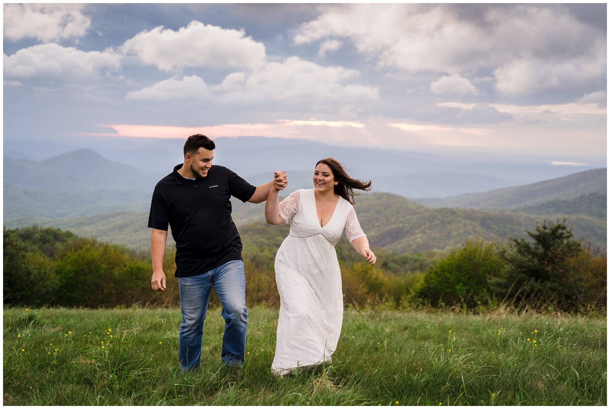 young engaged couple holding hands walking through grassy field overlooking mountain range smiling at one another