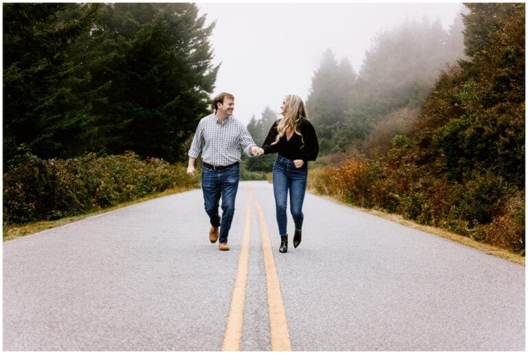 young engaged couple holding hands smiling at one another while running down the middle of a forested road for their outdoor winter engagement photo session