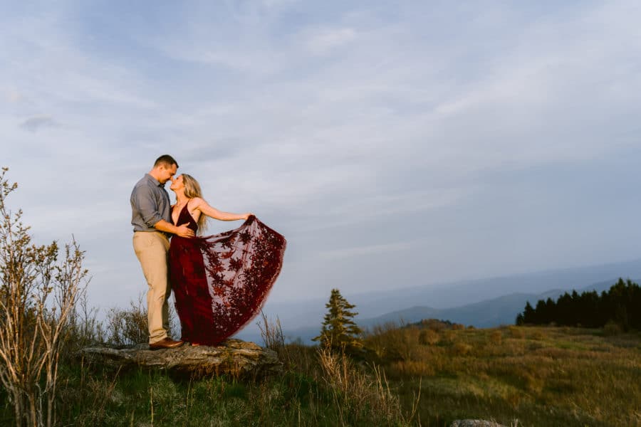 Engaged couple holding hands smiling at one another standing on rock surrounded by green trees and distant mountain views near Asheville, North Carolina photography done by Kathy Beaver.