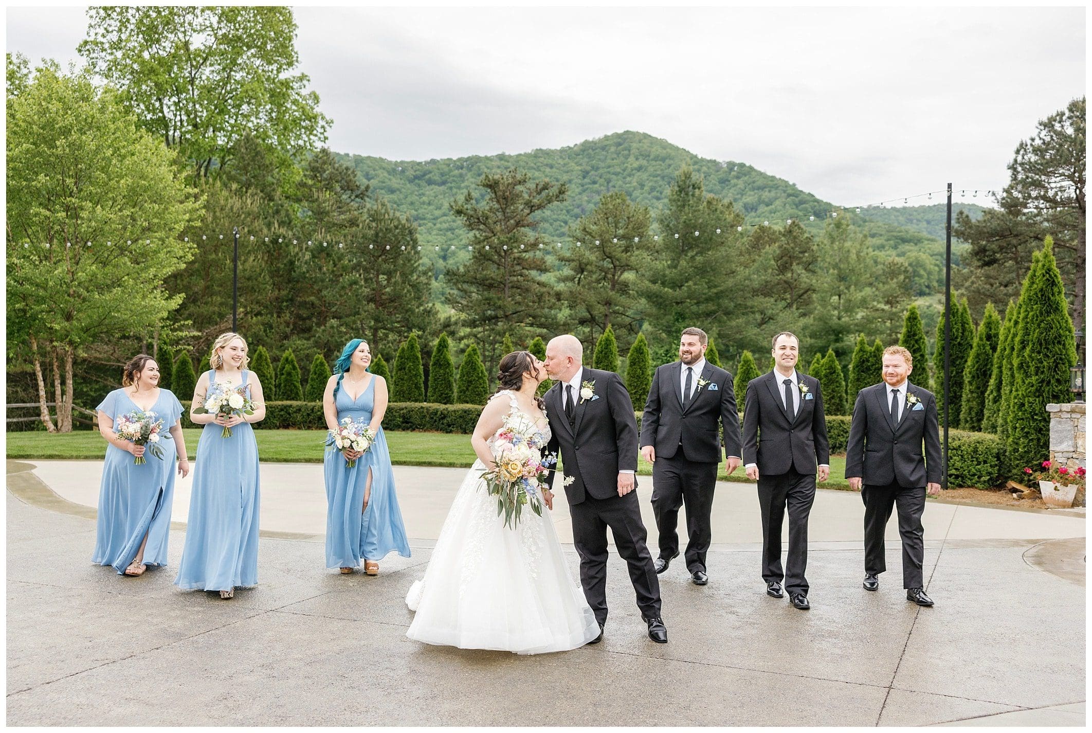 wedding party walks together with the bride and groom kissing at spring wedding with blue accents