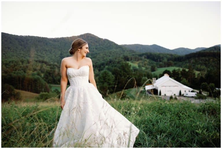 An elegant bride in a white wedding dress standing in a field with mountains as the backdrop for a summer wedding in Asheville.