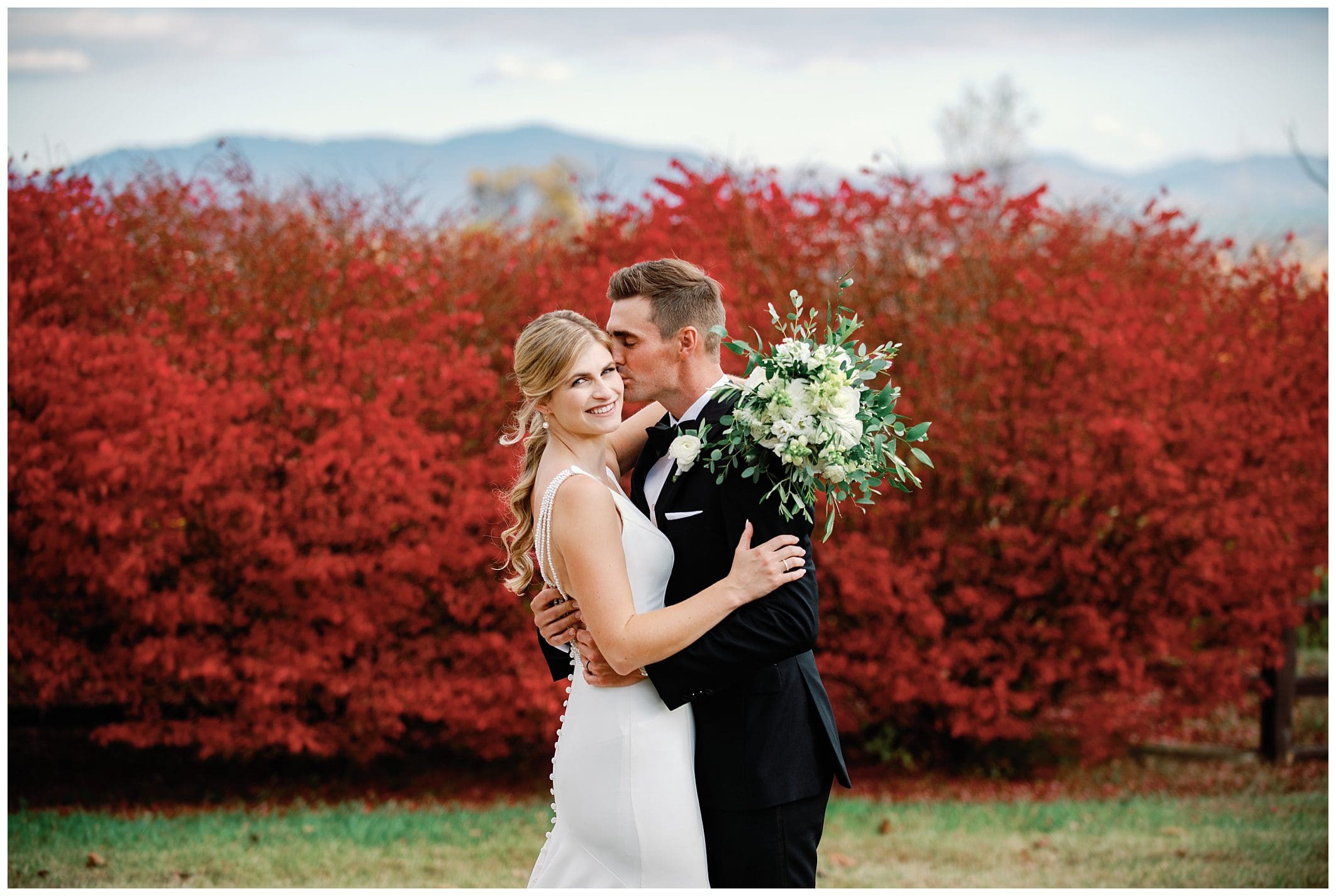 A bride and groom embrace in front of red bushes during their fall wedding at the Crest Center.