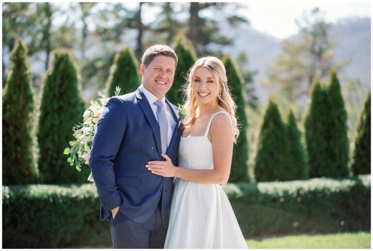 A bride and groom posing for a photo in front of bushes.