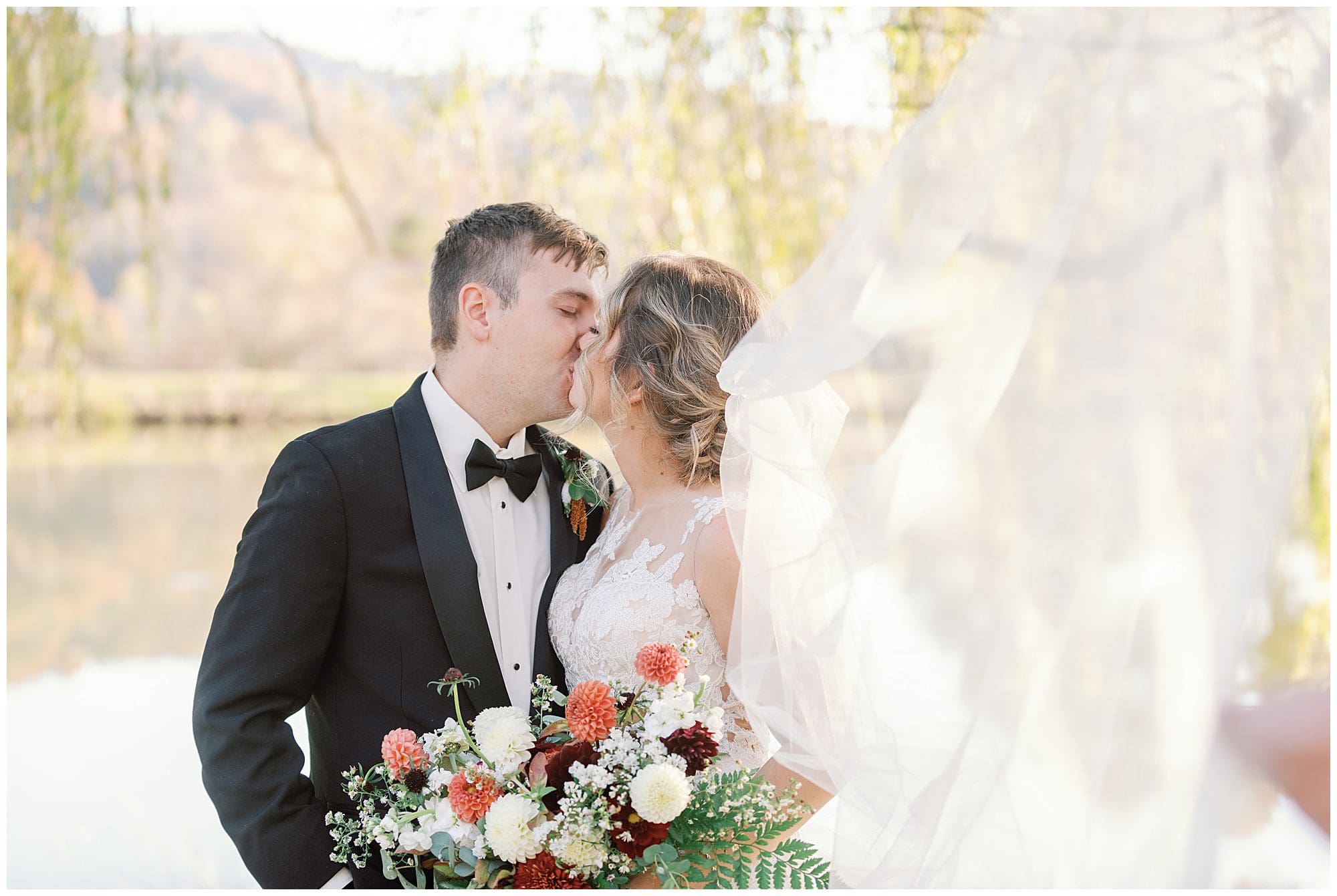 A bride and groom kissing outdoors with the bride's veil flowing in the air, holding a bouquet, with a tranquil lake and trees in the background.