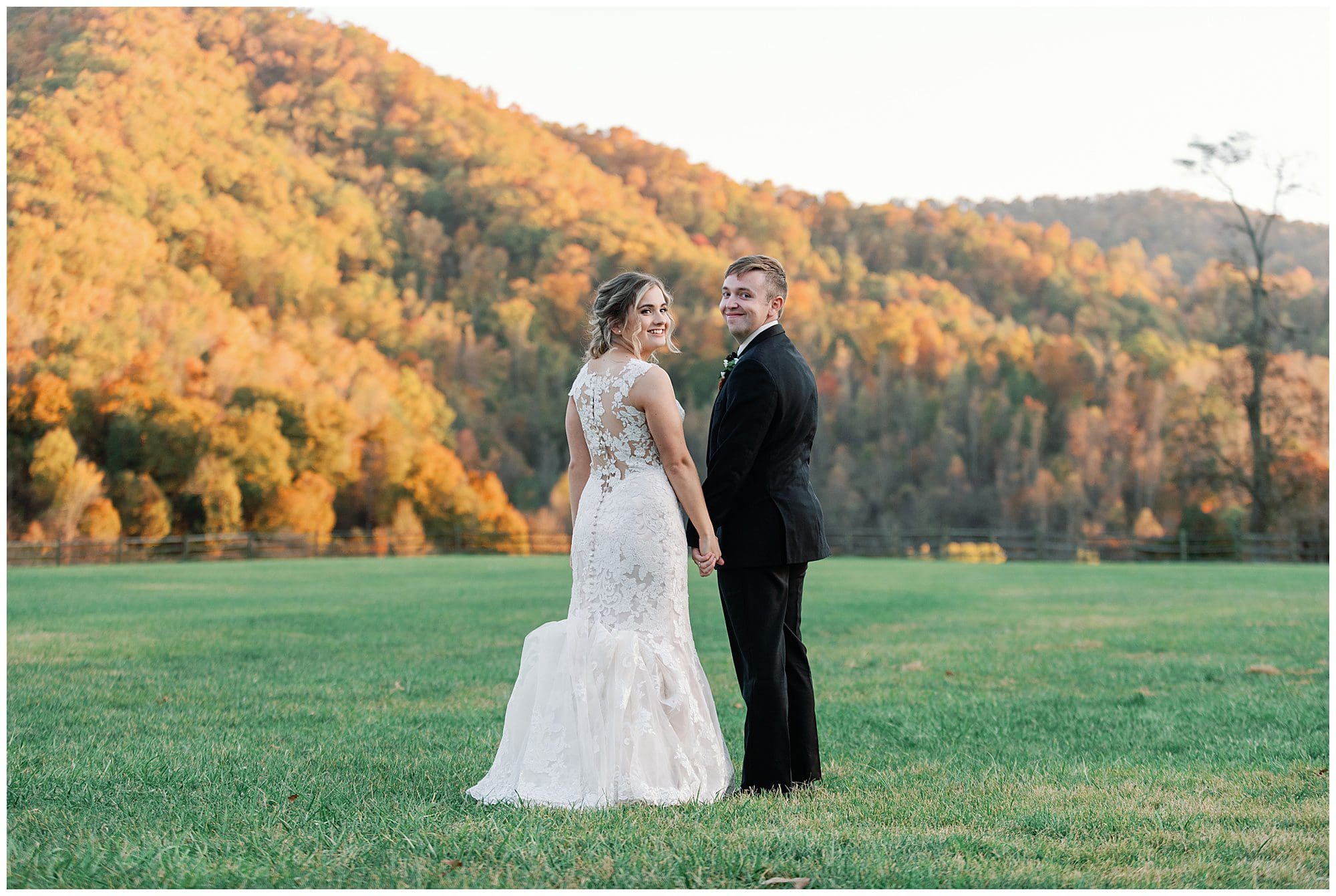 A bride and groom smile while holding hands in a field, with autumn-colored trees in the background.