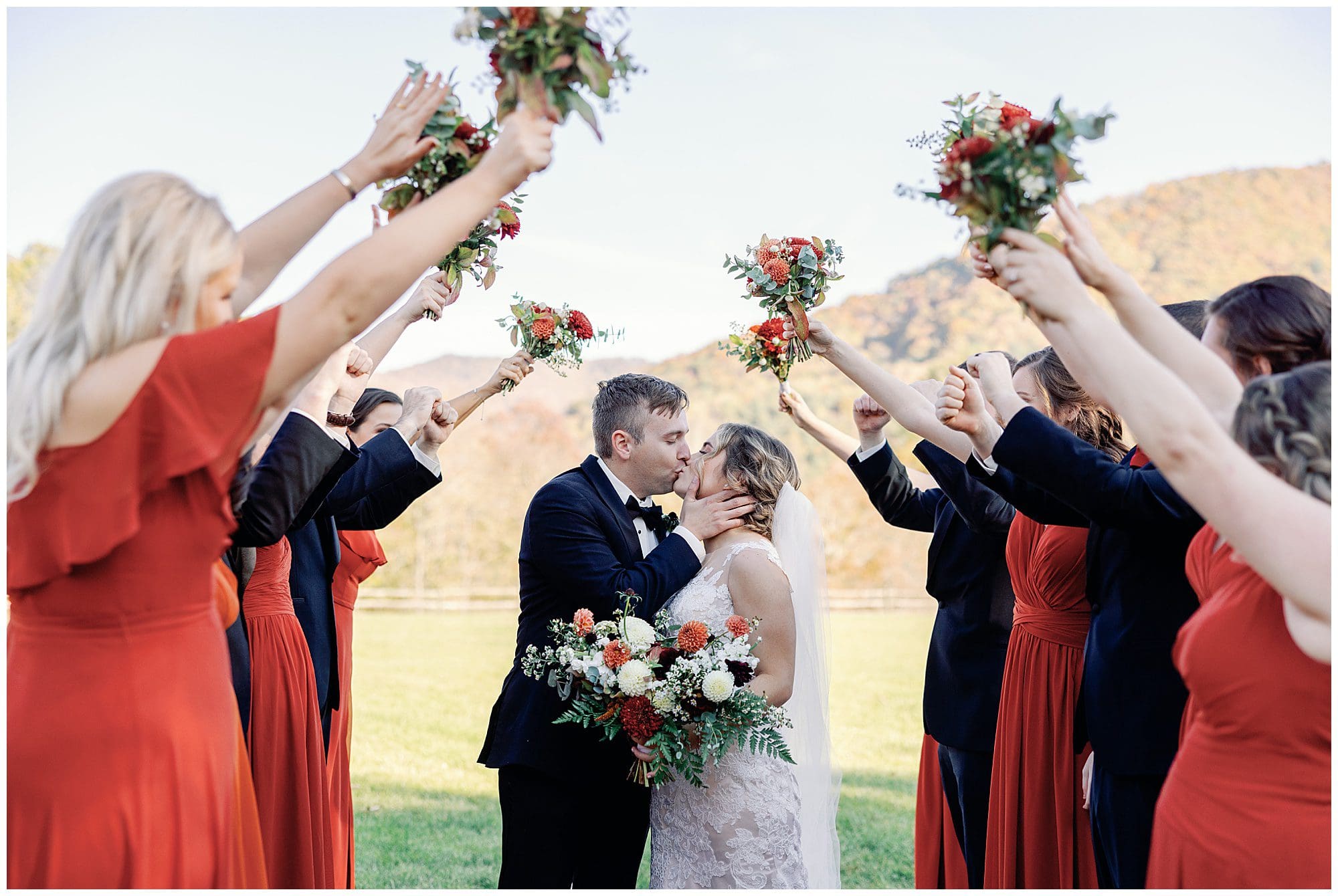 Bride and groom kissing under an arch formed by guests' raised arms holding bouquets, with a scenic mountain backdrop.