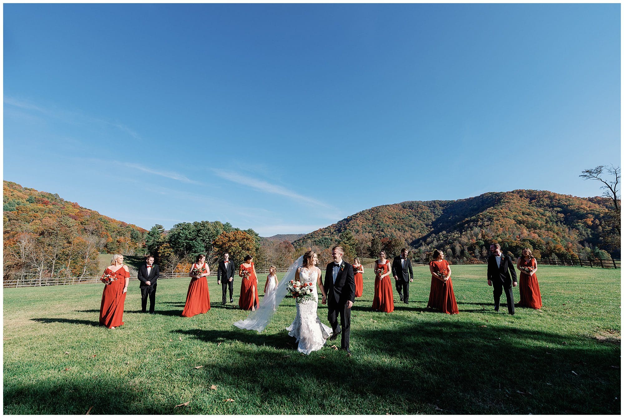 A wedding party outdoors with a bride and groom walking forward, surrounded by bridesmaids in red dresses and groomsmen in black suits, set against a scenic mountain backdrop.