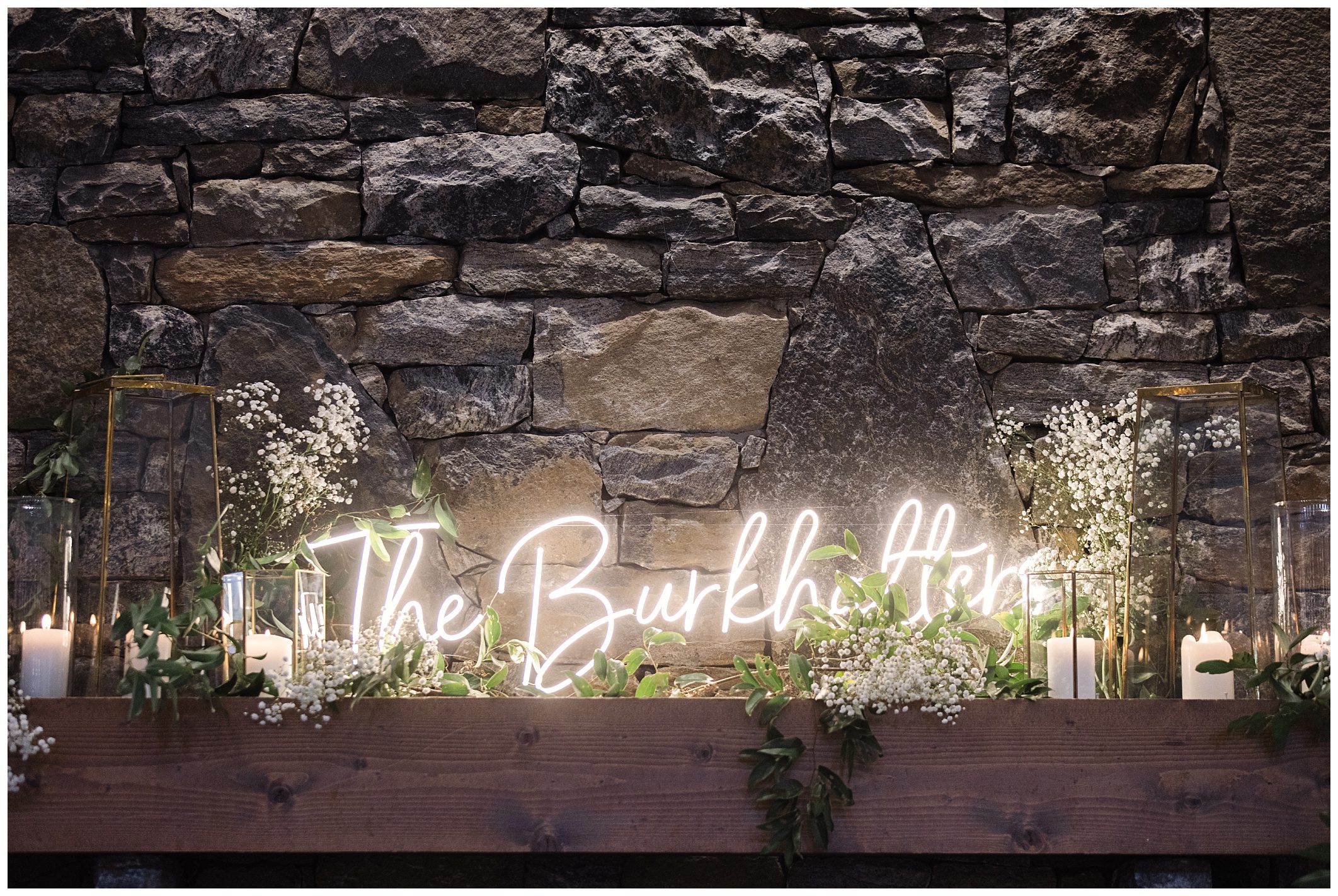 Decorative setup with the neon sign "the burkett tiek" on a stone wall at Chestnut Ridge, flanked by white flowers and candles in glass holders.