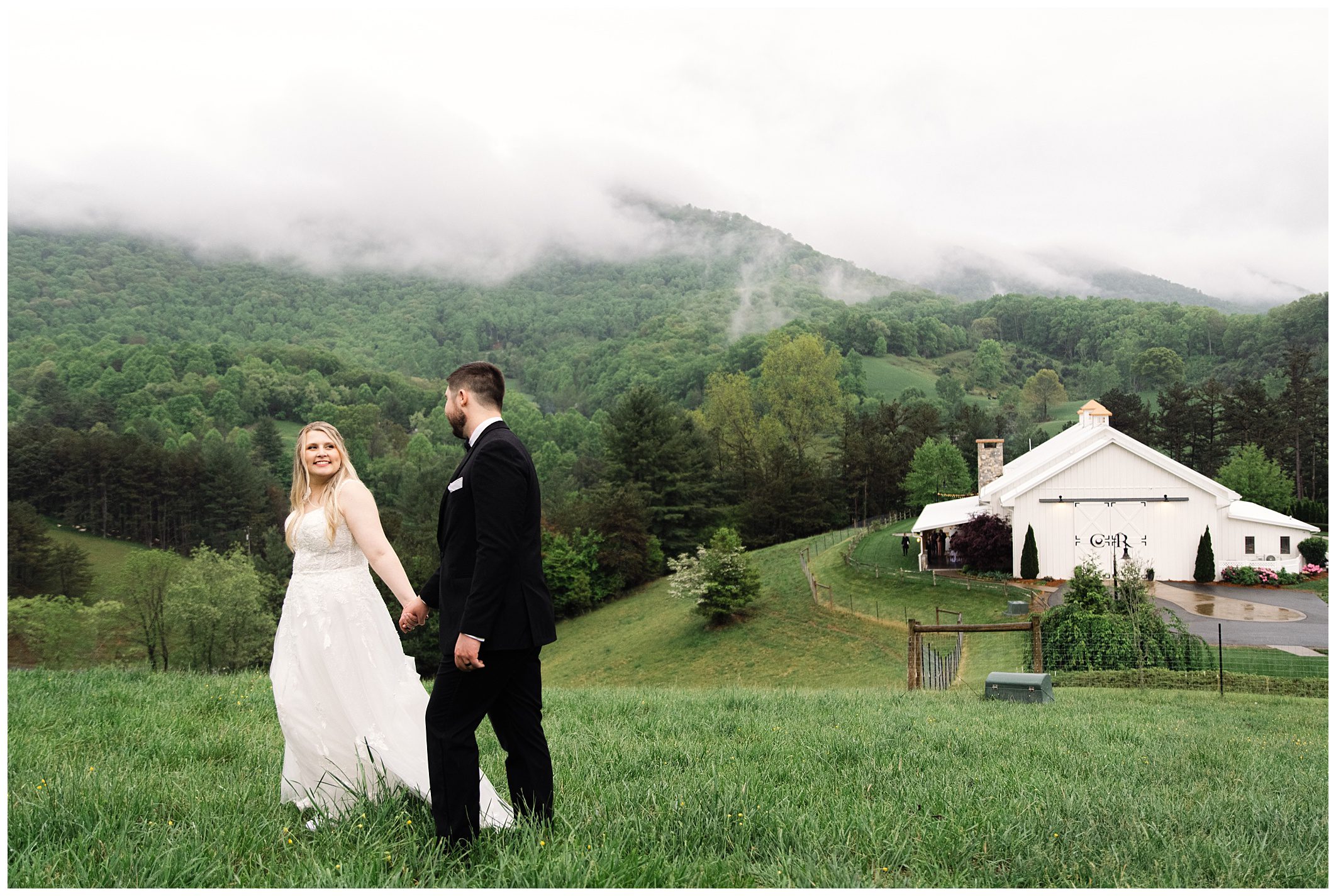 A bride in a white dress and a groom in a black suit holding hands and smiling at each other on a green hill with foggy mountains and rain at Chestnut Ridge in the background.