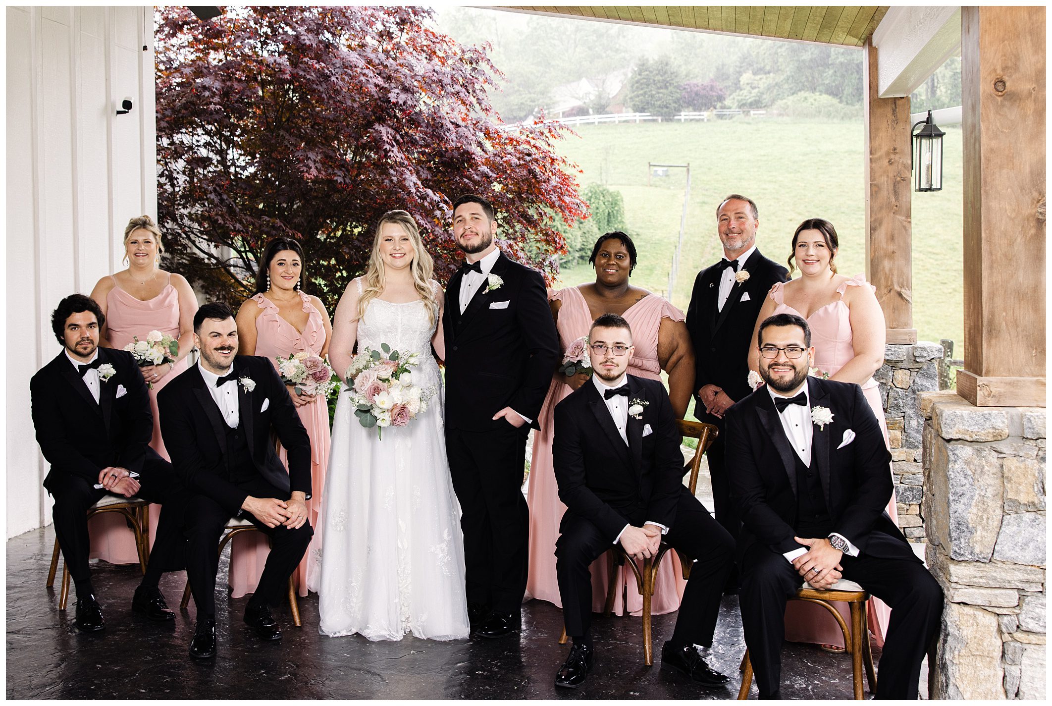 Wedding party posing together under a covered patio at Chestnut Ridge, featuring a bride and groom in the center, with bridesmaids in pink and groomsmen in black tuxedos.