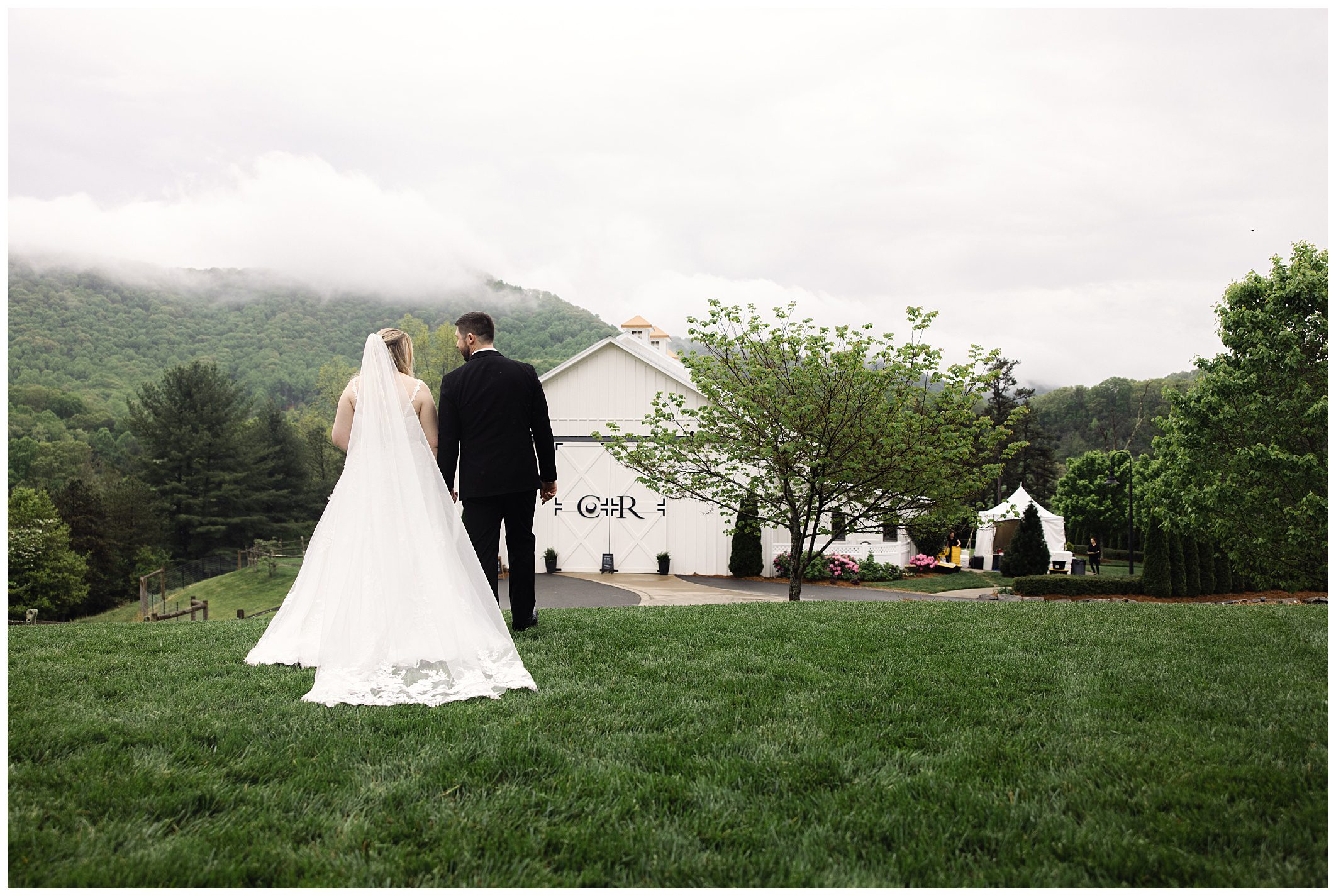 A bride and groom holding hands, walking towards a white chapel with mountains in the background on a rainy day at Chestnut Ridge.