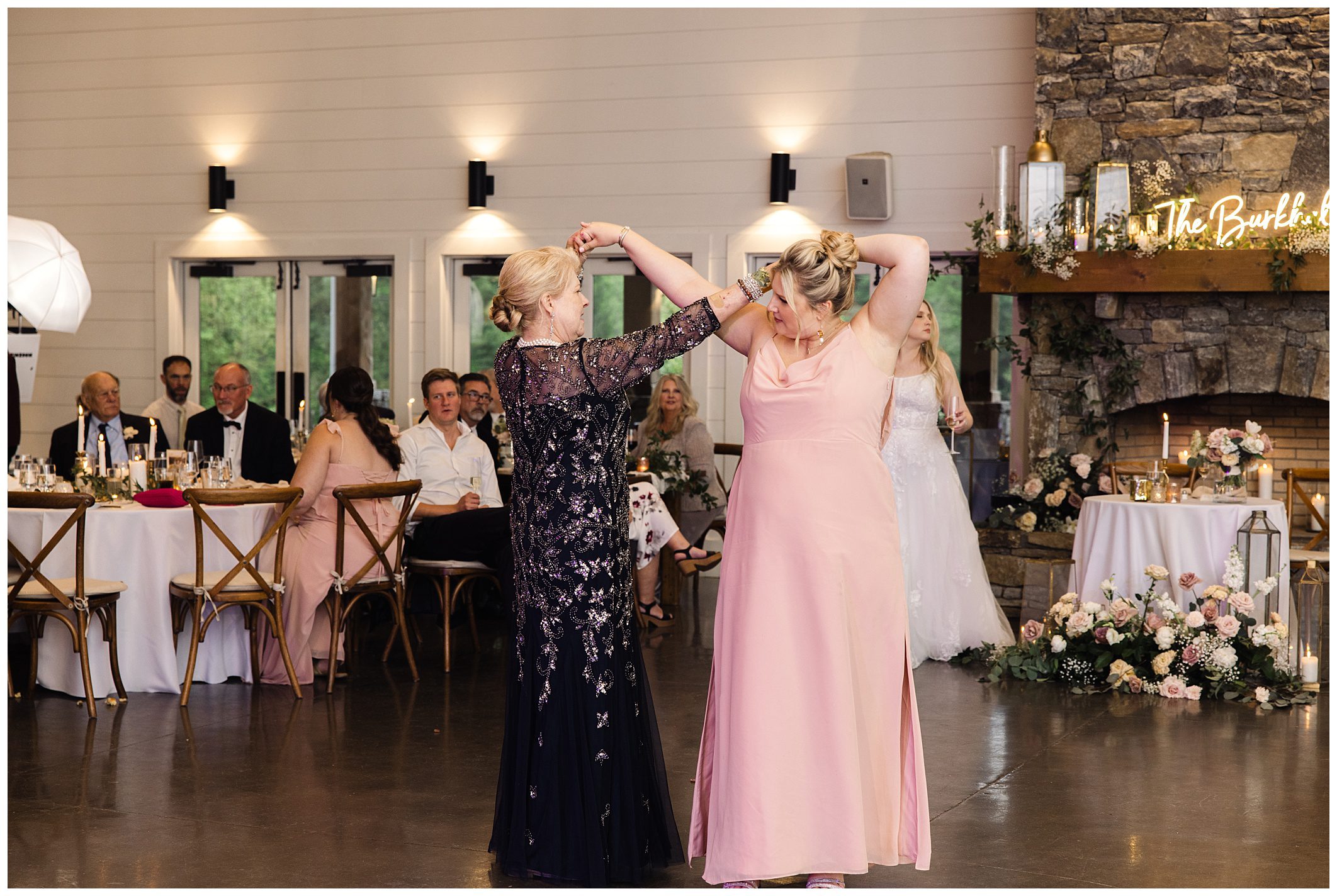 Two women dancing joyfully together at a mountain wedding reception, with guests seated at tables in the background.