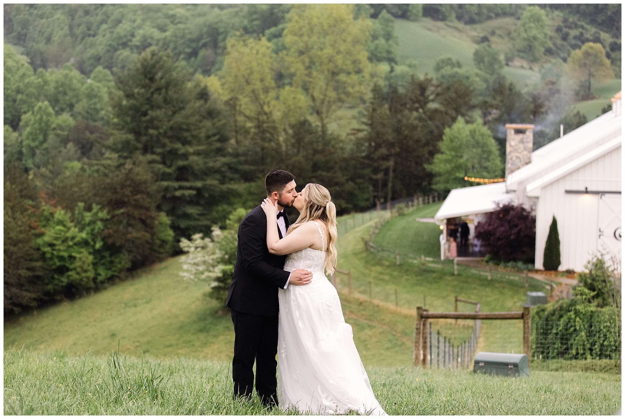 A bride and groom kiss in a scenic outdoor setting with green hills and a quaint building in the background during a mountain wedding at Chestnut Ridge.