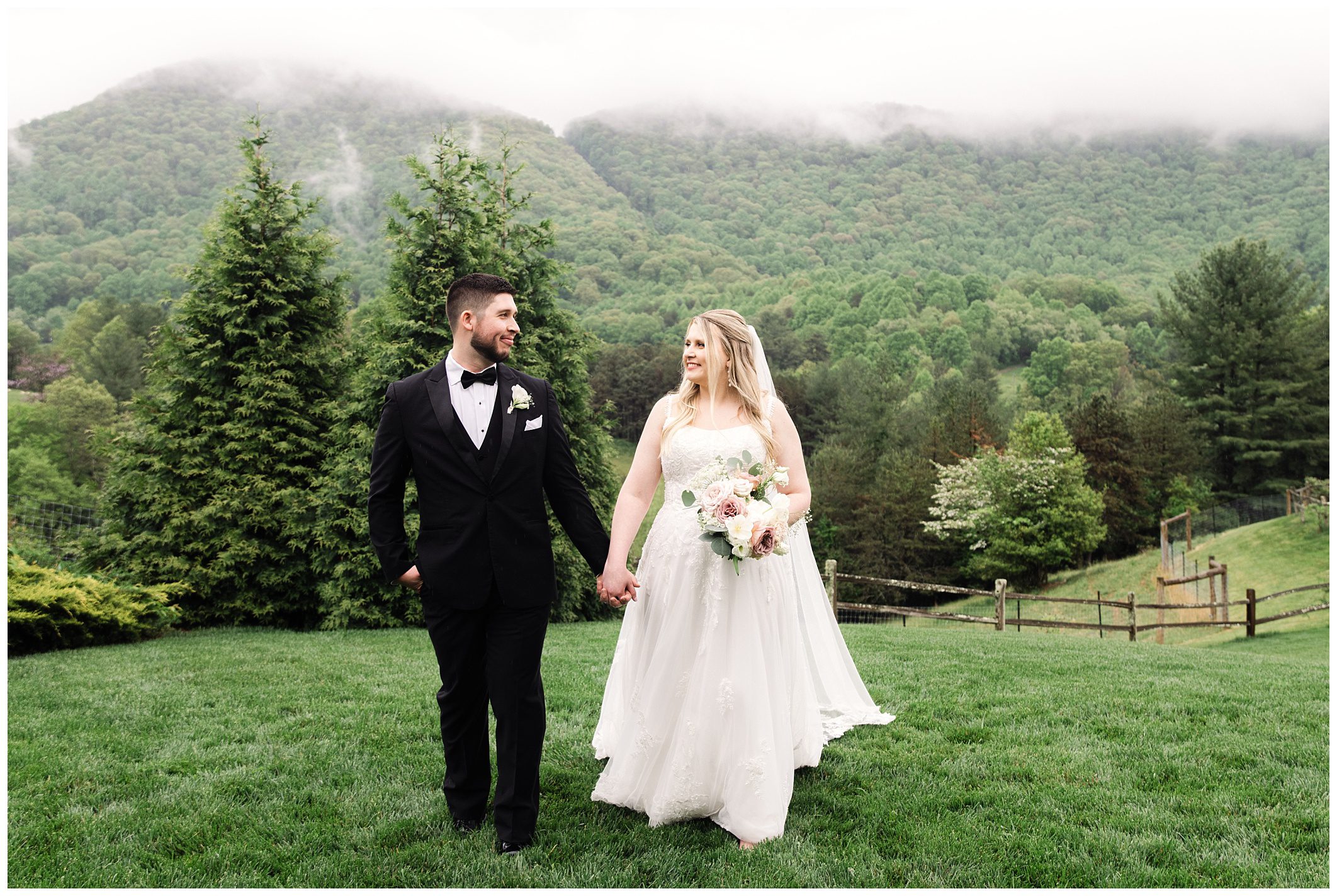 A bride and groom holding hands while walking across a grassy field at Chestnut Ridge, with mist-covered mountains in the background.