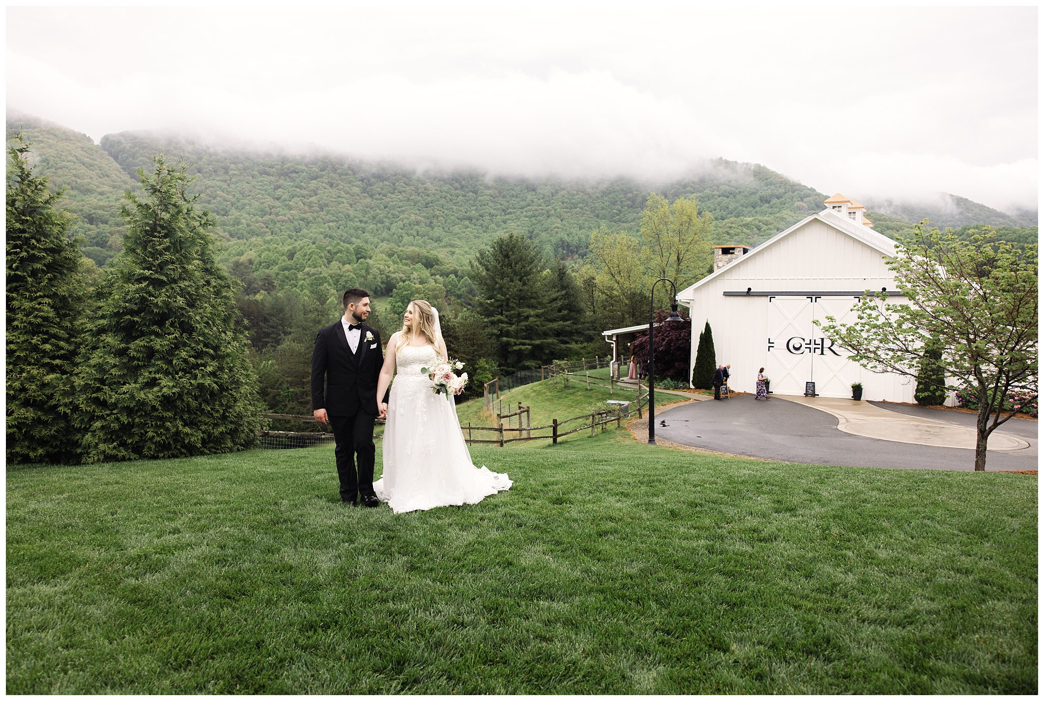 Bride and groom holding hands in a grassy field, with a white barn and misty mountains at Chestnut Ridge in the background.