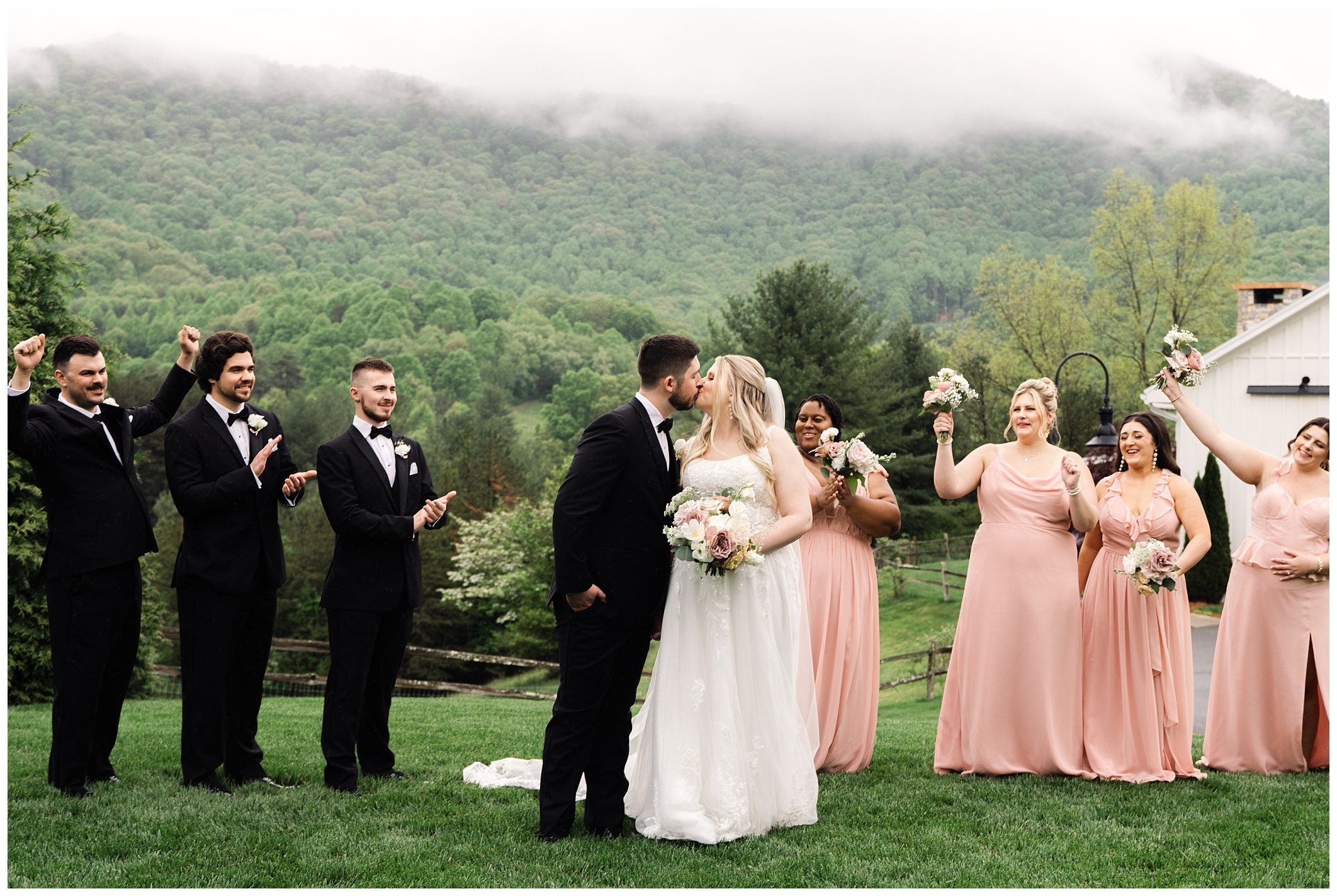 Bride and groom kissing in front of a wedding party, with rain-soaked mountains at Chestnut Ridge in the background and guests wearing black and pink.