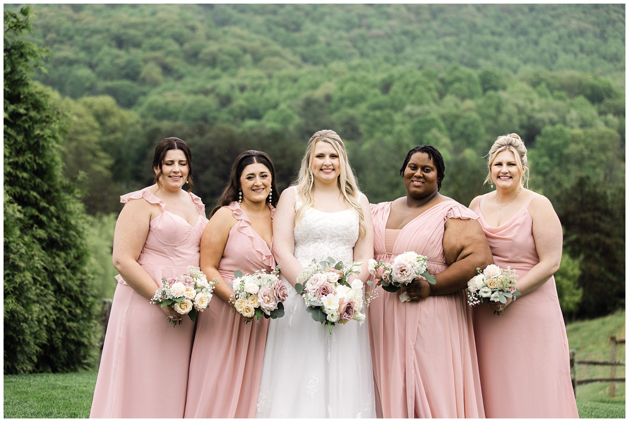 A bride in a white gown stands with four bridesmaids in pale pink dresses, all holding bouquets, with green mountains at Chestnut Ridge in the background.