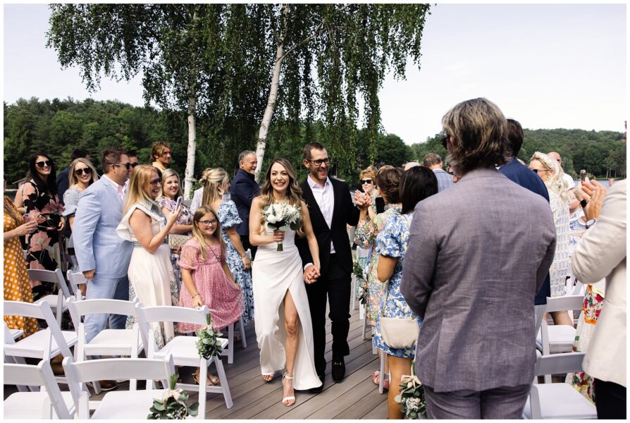 A bride and groom walk down an outdoor aisle surrounded by applauding guests. The bride holds a bouquet, and the groom wears a dark suit. There are trees in the background.