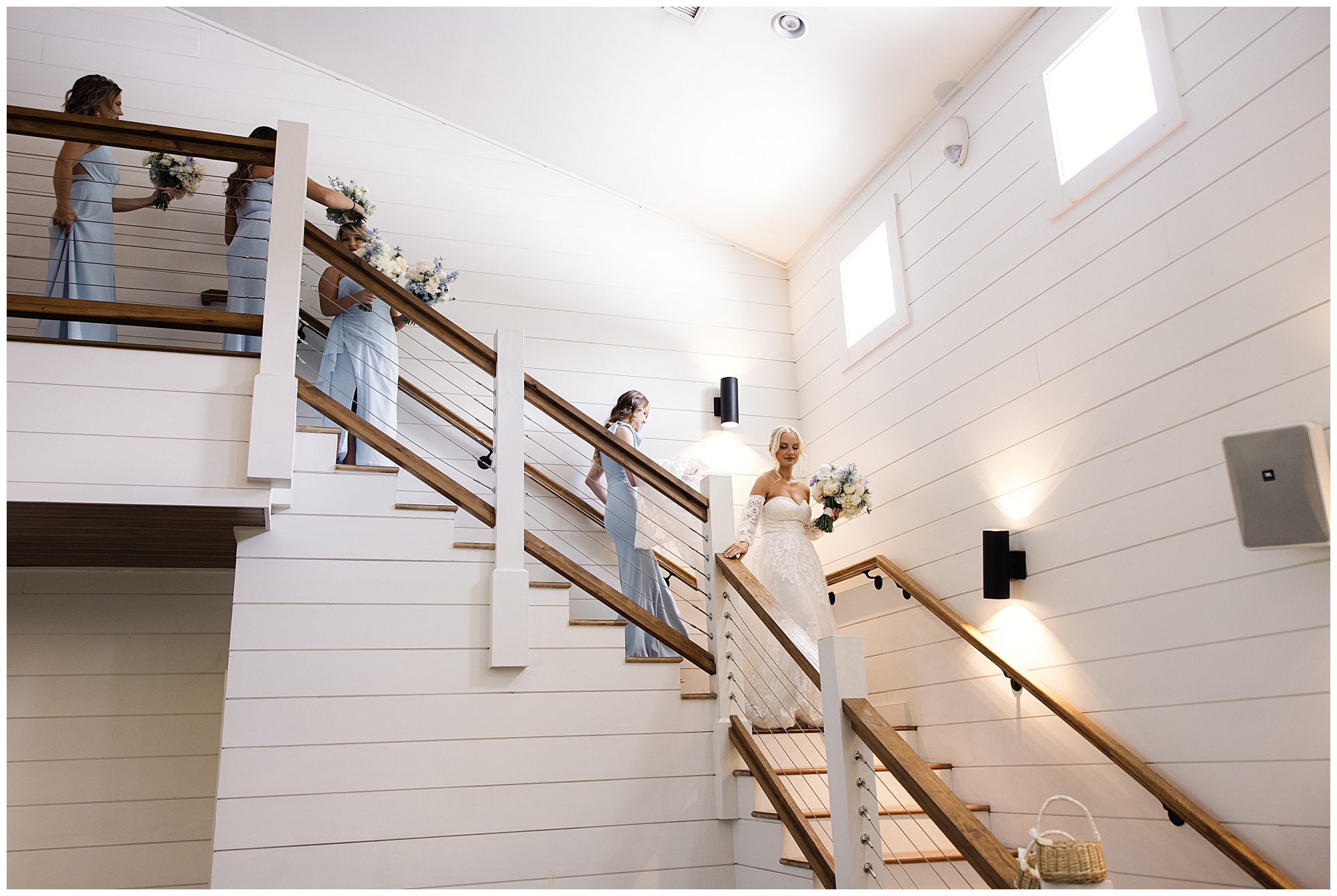 A bride descends a staircase in a white dress, holding a bouquet. Three bridesmaids in light blue dresses follow, each carrying bouquets. The interior is modern with white walls and wooden railings.