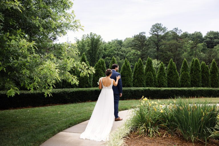 A bride and groom stand together on a path in a garden during their summer wedding in Asheville. The groom faces away while the bride touches his shoulder, surrounded by lush greenery and trees.
