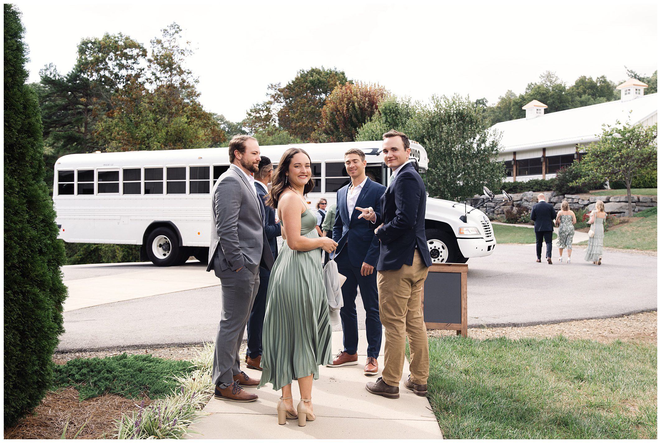 A group of people in formal attire stand outdoors near a white bus, with a few smiling and looking at the camera. Trees and a building are in the background.