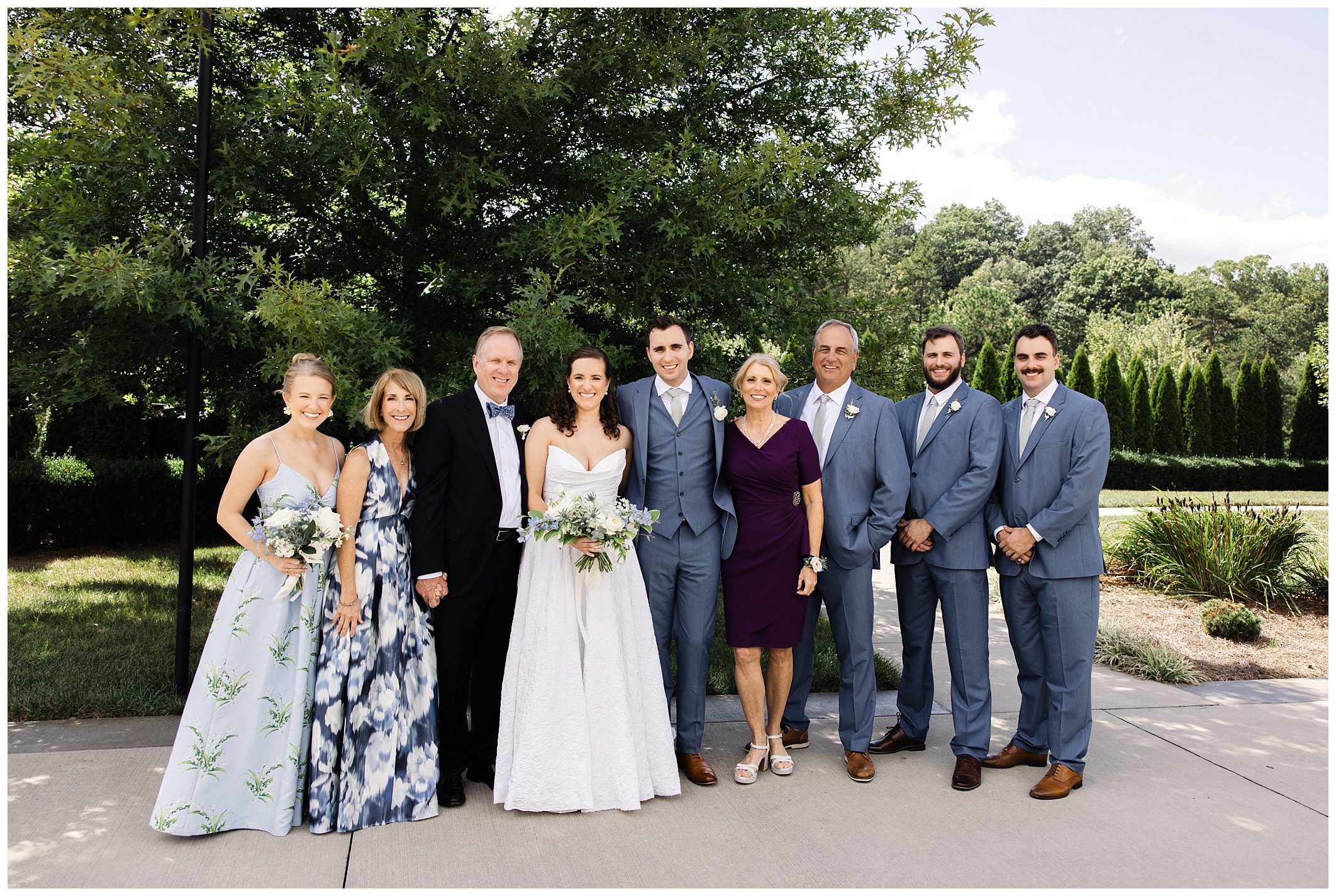 A wedding party stands outdoors, featuring the bride and groom in the center, flanked by family and friends. The groom's party wears blue suits, and the bride's party wears various shades of blue dresses.