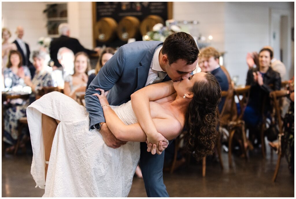 A couple in wedding attire shares a kiss while dancing as guests seated at tables look on in the background.