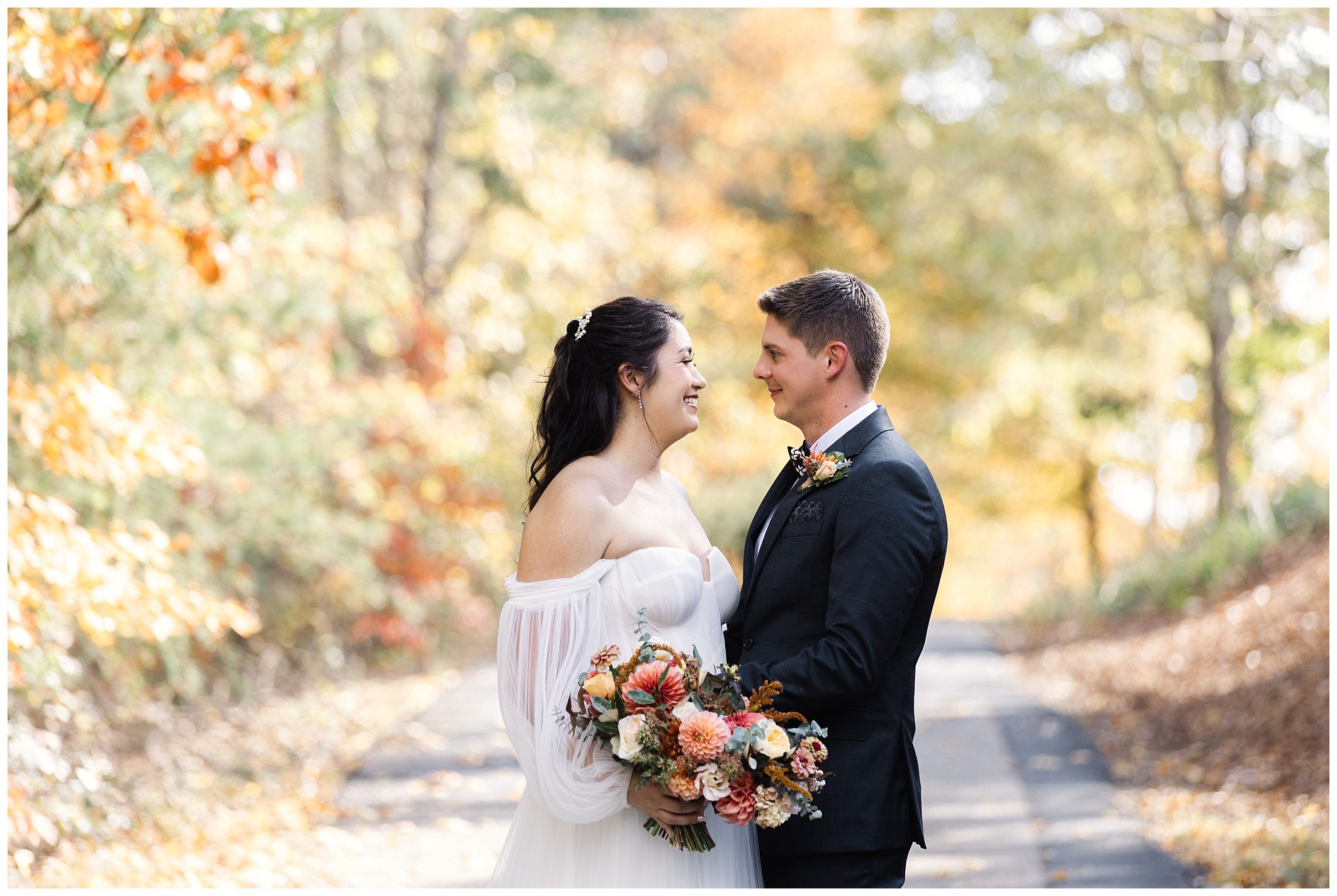 Bride and groom smiling at each other on a tree-lined path, surrounded by autumn foliage.