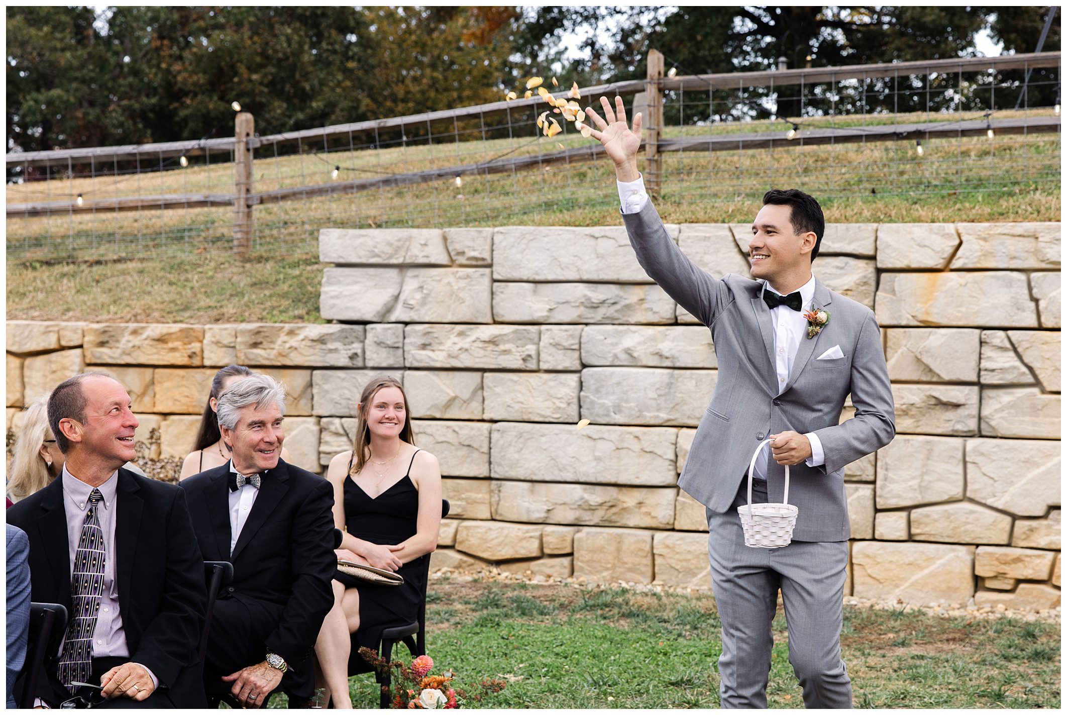 A man in a gray suit tosses petals while walking down an outdoor aisle lined with seated guests.