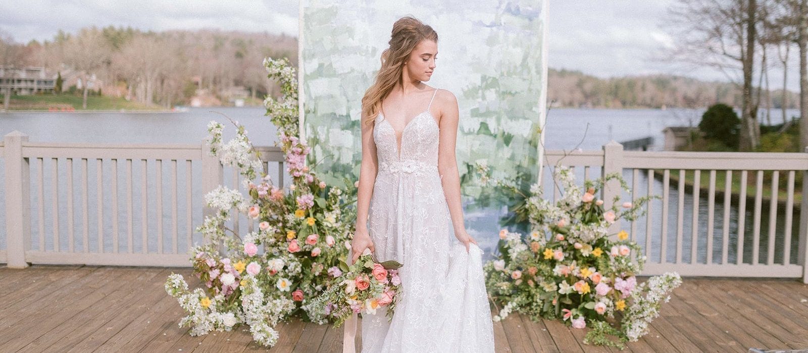 Bride holding bouquet down by side and looking down to the left standing in front of white, green and blue painted canvas backdrop on deck over looking a lake