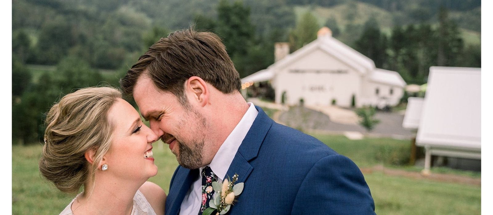 Bride and groom standing in field overlooking blue ridge mountains hugging and smiling at one another about to kiss