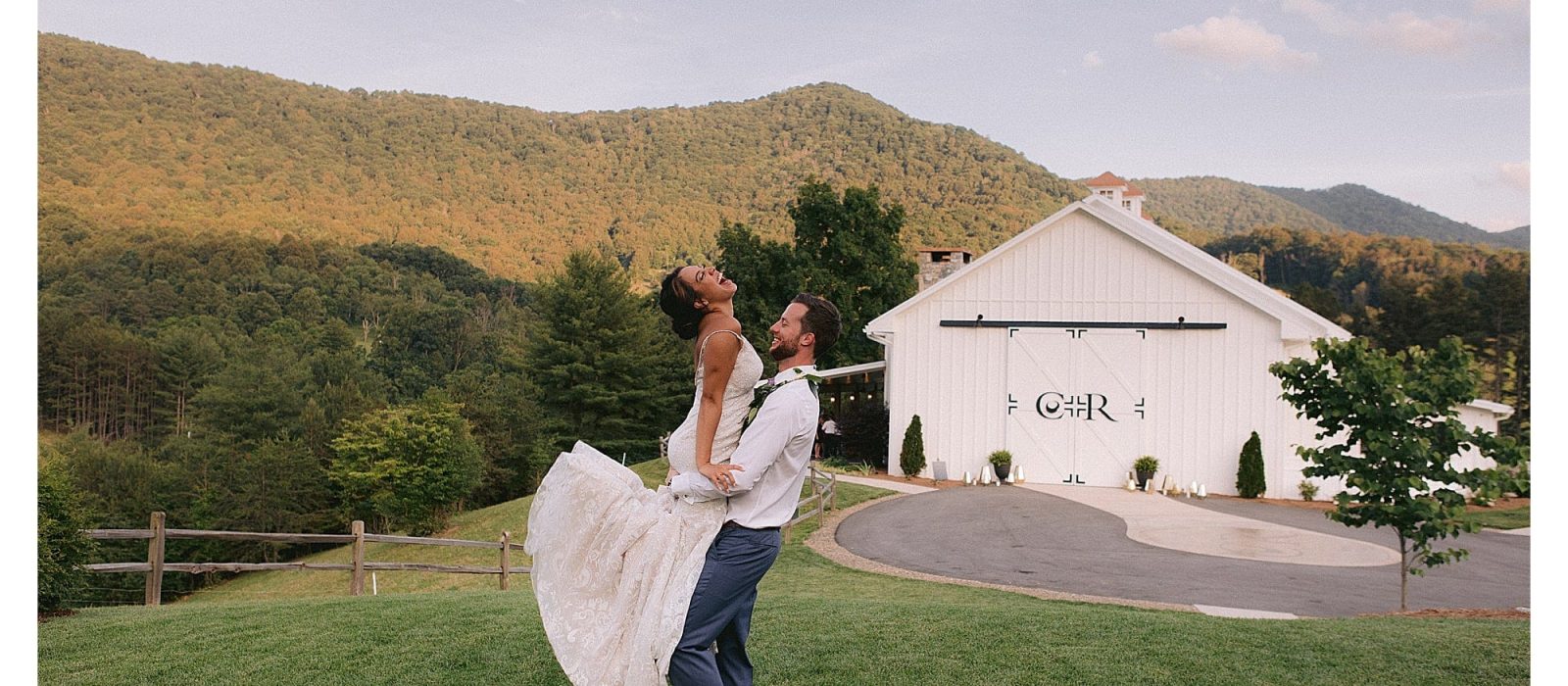 groom hugging bride and lifting her up in grassy field with mountains in background