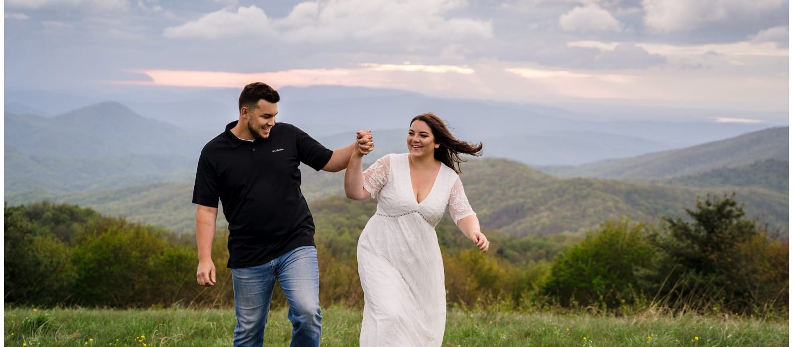 young engaged couple holding hands walking through grassy field overlooking mountain range smiling at one another