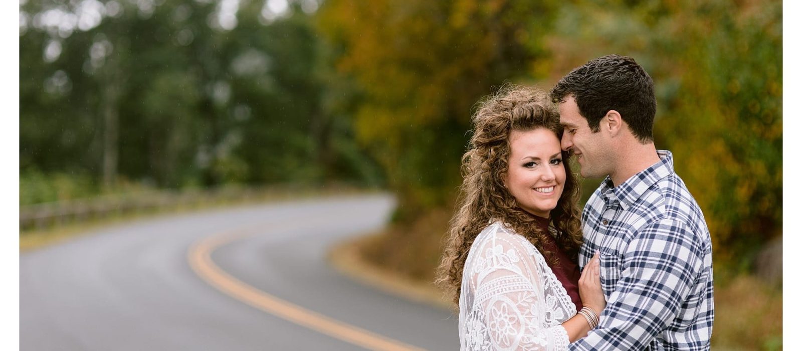 Engaged couple standing on road hugging one another and smiling with autumn trees behind them