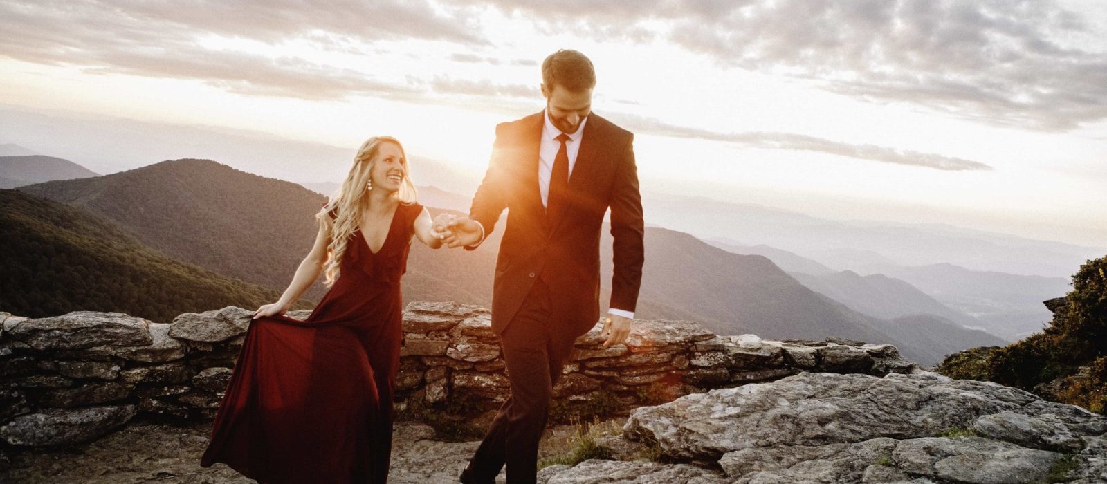 Man in suit holding fiances hand walking along rocky mountain side with mountain range in background at sunset