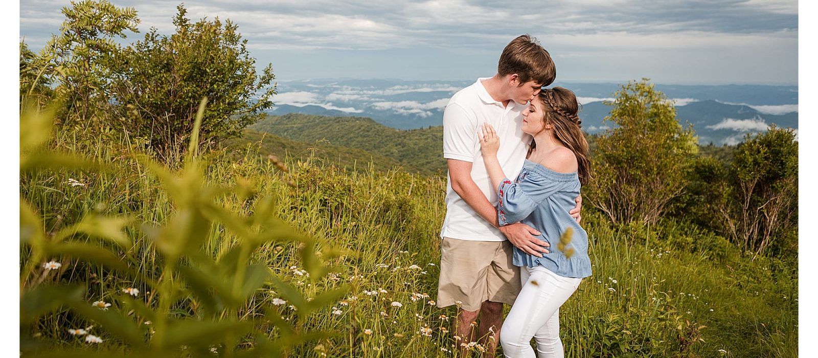Young man kissing fiances forehead while standing in field with mountains in background